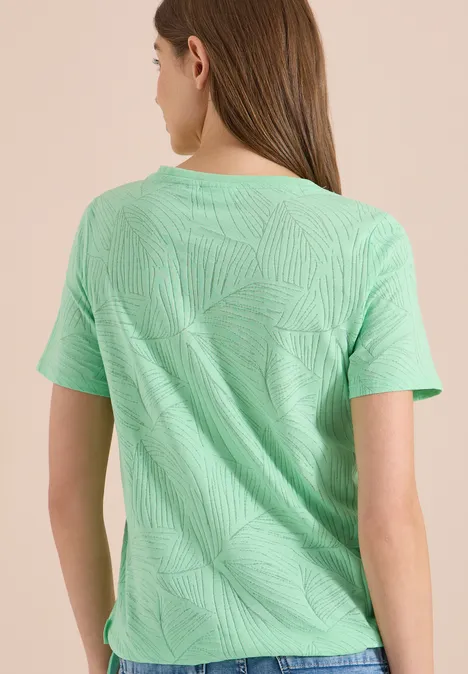 Frau steht mit dem Rücken zur Kamera in mintgrünem T-Shirt mit dezentem Blattmuster vor beigem Hintergrund.