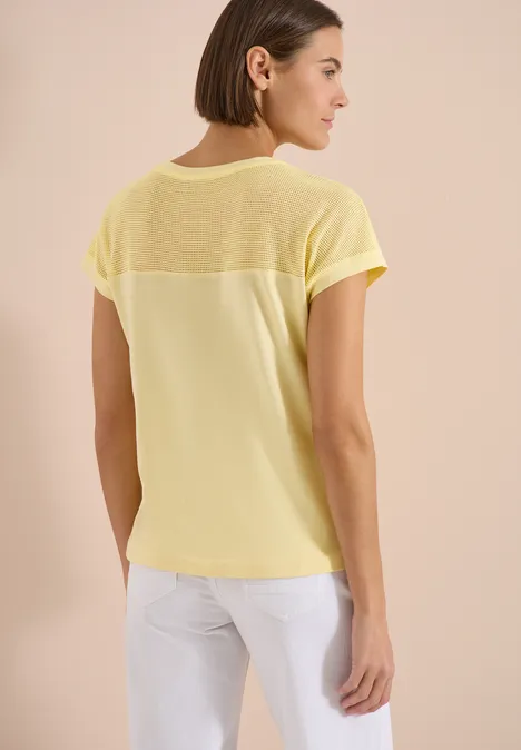 Vrouw van achteren in een losvallend geel T-shirt met transparante inzet op de rug.