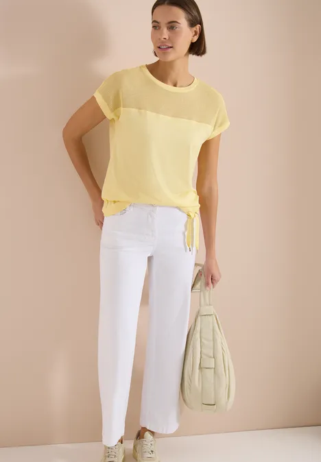 Model draagt transparant geel kort shirt met knoop, witte cropped broek en een pastelgroene tas.