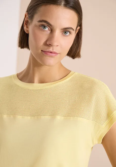 Vrouw in pastelgeel, fijngebreid topje met korte mouwen tegen een lichte, neutrale achtergrond.