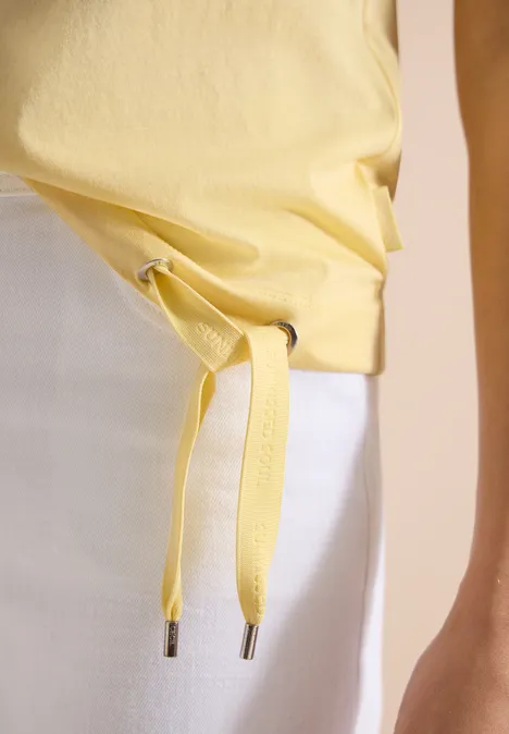 Geel T-shirt aan de taille geknoopt, gecombineerd met een witte rok en een arm in beeld.
