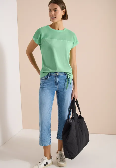 Vrouw in lichtgroene top met knoop, blauwe 7/8-jeans, grijze sneakers en zwarte shopper.