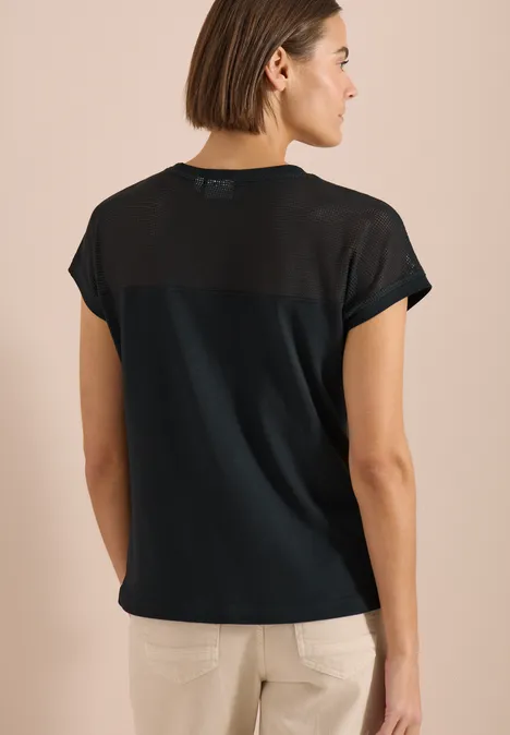 Achteraanzicht van een vrouw in een zwart T-shirt met transparante rugpartij en lichte broek.