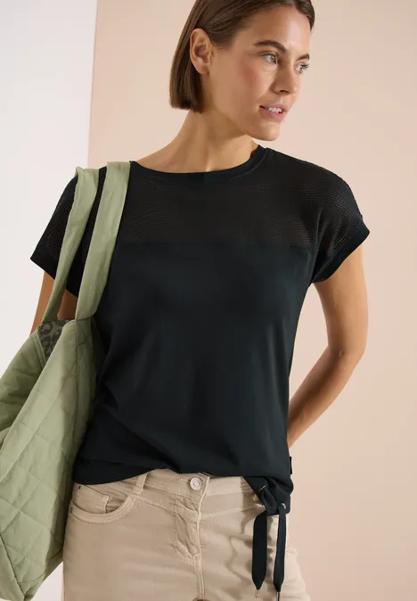 Vrouw draagt donkerblauw T-shirt met transparante inzet bij de schouders, knoop onderaan en groene tas.