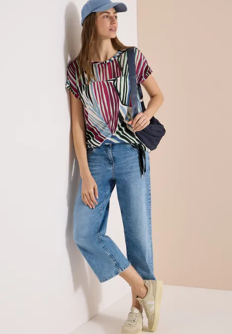 Frau lehnt an Wand, trägt bunte Streifenbluse, weite Jeans, Cap, Umhängetasche und helle Sneaker.