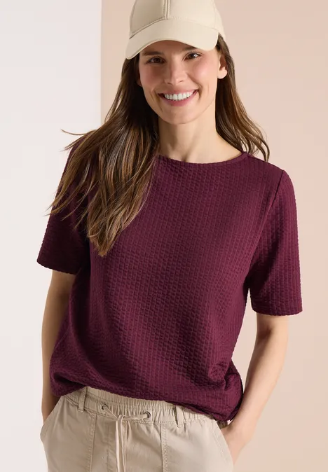 Vrouw in een gestructureerd paarse t-shirt en beige broek, met één hand in de zak.