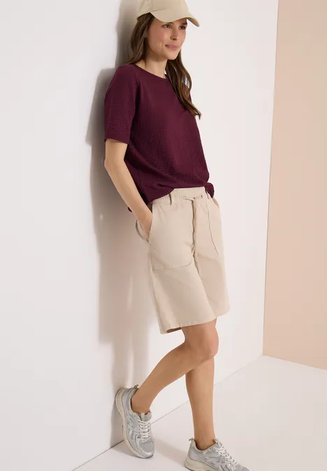 Een persoon leunt tegen een muur, draagt een bordeauxrood t-shirt, beige shorts en grijze sneakers.