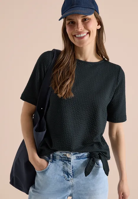 Frau mit schwarzem, geknotetem Kurzarmshirt, blauer Jeans, Cap und Schultertasche vor neutralem Hintergrund.