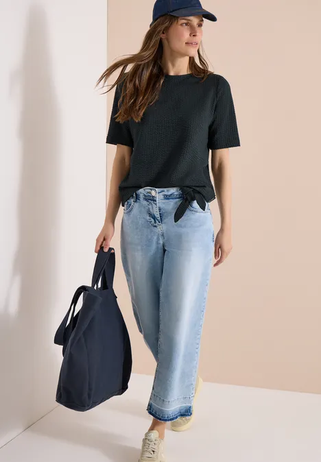 Frau in schwarzem T-Shirt, Jeans und Cap trägt eine große blaue Tasche.