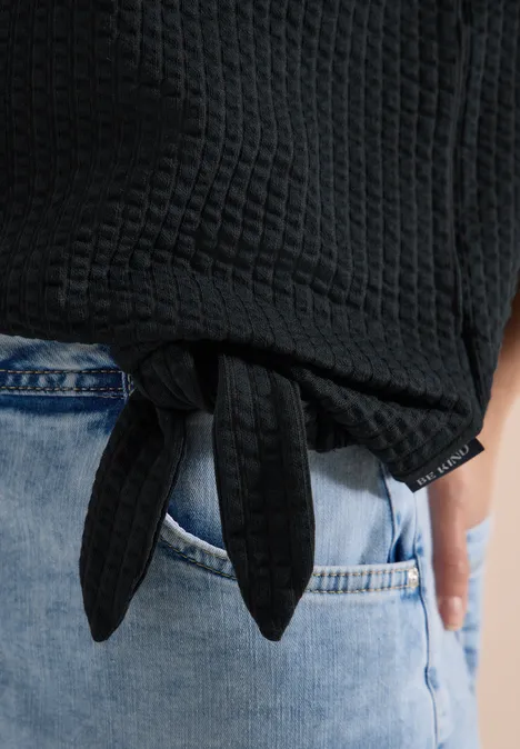 Schwarz-texturierter Pullover mit Knotendetail am Bund, kombiniert mit hellblauen Jeans.