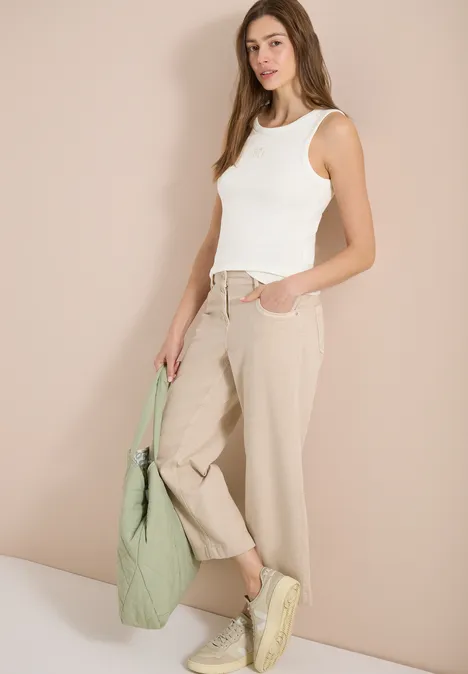 Frau trägt weißes Top, beige Hose, gelbe Sneakers und hält eine grüne Tasche.