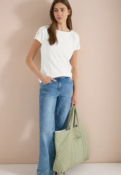 Vrouw in wit T-shirt en blauwe jeans met een doorgestikte groene tas voor een beige achtergrond.