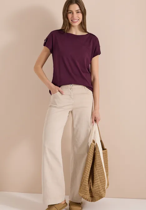 Vrouw in paars T-shirt, witte wijde broek en rieten tas.