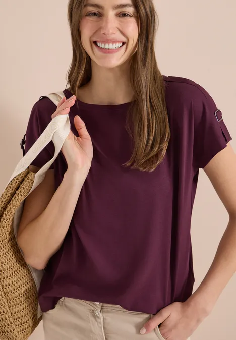 Vrouw in paarse T-shirt met korte mouwen en beige broek, met strooien tas over schouder.