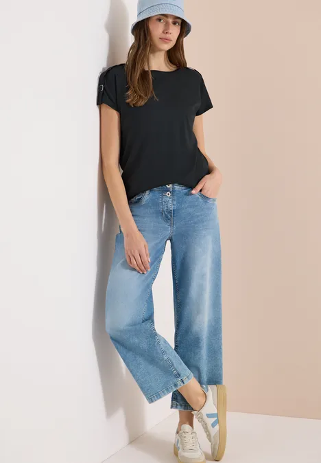 Frau in schwarzem T-Shirt, weiten blauen Jeans und weißen Sneakern vor zweifarbigem Hintergrund.