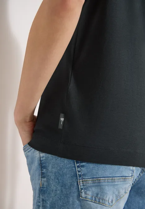 Eine Person trägt ein schwarzes T-Shirt und blaue Jeans, Hand in Hosentasche.