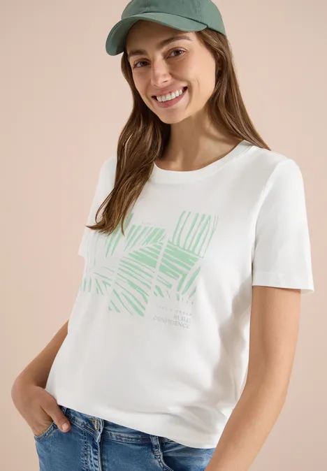 Frau mit weißem T-Shirt mit grünem Blattmuster, Jeans und blauer Kappe vor neutralem Hintergrund.