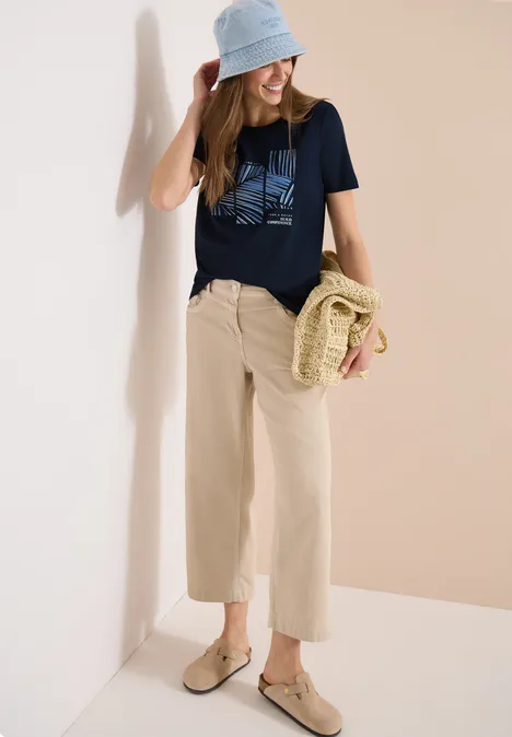 Frau in beiger Culotte, schwarzem T-Shirt, blauem Bucket Hat und Basttasche vor hellem Hintergrund.