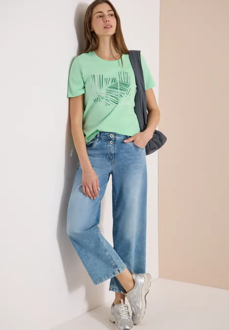 Frau lehnt an Wand, trägt grünes T-Shirt mit Print, weite Jeans-Culotte, weiße Sneaker und Denim-Tasche.