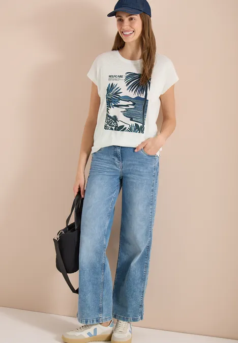 Jonge vrouw draagt wit t-shirt met palmenprint, wijde jeans, sneakers, pet en rugzak.