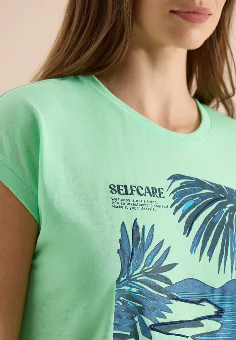 Vrouw draagt groen T-shirt met opschrift 'SELFCARE' en blauwe palmbladeren.