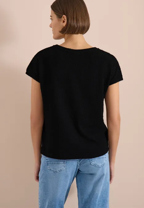 T-Shirt mit Boatneck im Ajour-Look Black Frau von hinten in schwarzem T-Shirt und hellblauen Jeans vor beigefarbenem Hintergrund.