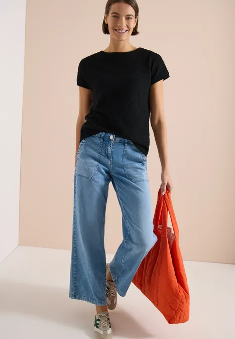 T-Shirt mit Boatneck im Ajour-Look Black Frau in schwarzem T-Shirt, weitem Jeans und orangefarbener Tasche vor hellem Hintergrund.