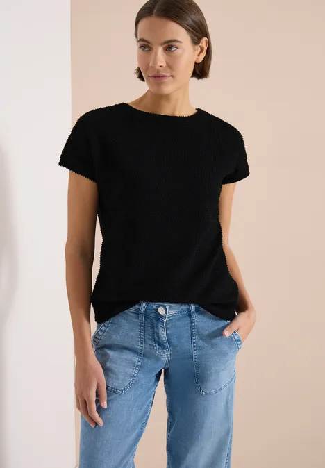 T-Shirt mit Boatneck im Ajour-Look Black Frau in schwarzem T-Shirt und blauen Jeans vor schlichtem, beigem Hintergrund.