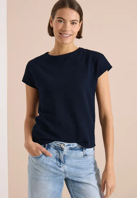 T-Shirt mit Boatneck im Ajour-Look universal blue Frau trägt dunkelblaues T-Shirt mit kurzen Ärmeln und helle Jeans vor beigem Hintergrund.