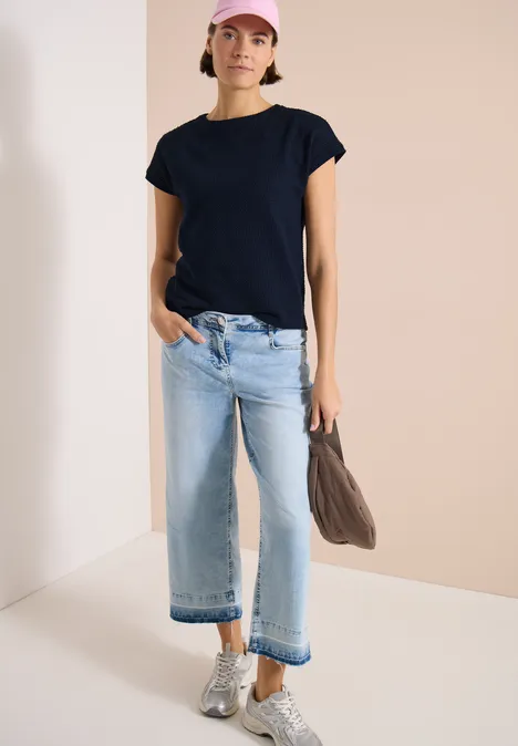 T-Shirt mit Boatneck im Ajour-Look universal blue Frau in schwarzem T-Shirt, hellblauen Kickflare-Jeans, silbernen Sneakern und Taupe-Umhängetasche.