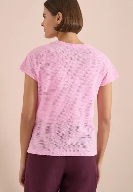 Eine Frau von hinten in rosa Kurzarmshirt und bordeauxroter Hose vor hellem Hintergrund.