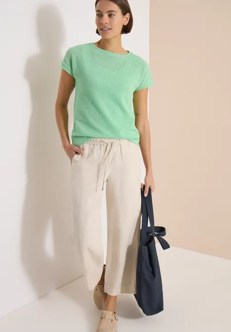 T-Shirt mit Boatneck im Ajour-Look juicy lime Frau in grünem T-Shirt und weißer, weiter Hose mit Schlitz, trägt eine blaue Tasche vor hellem Hintergrund