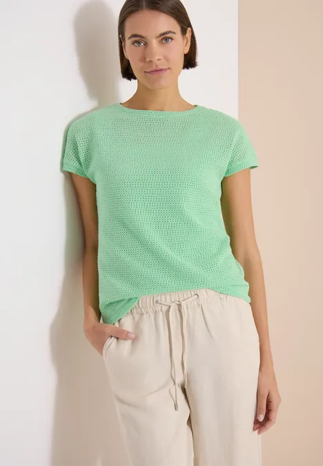 T-Shirt mit Boatneck im Ajour-Look juicy lime Frau in grüner Kurzarmbluse und hellen Leinenhosen, Hand in Hosentasche vor schlichtem Hintergrund.