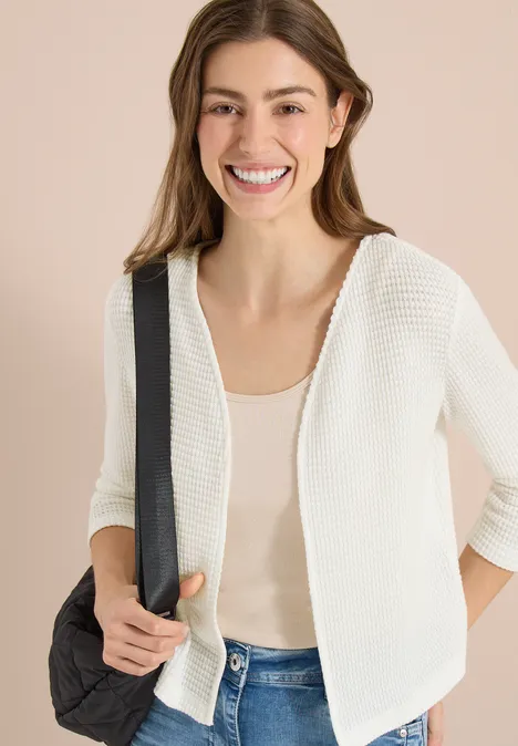 Vrouw draagt witte cardigan, beige top en jeans, houdt zwarte schouderriem vast