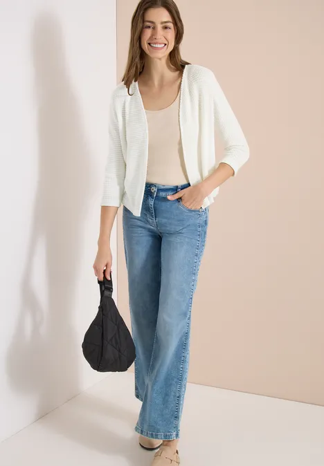 Vrouw in beige top, wit vest en blauwe jeans, met zwarte tas voor een beige muur.