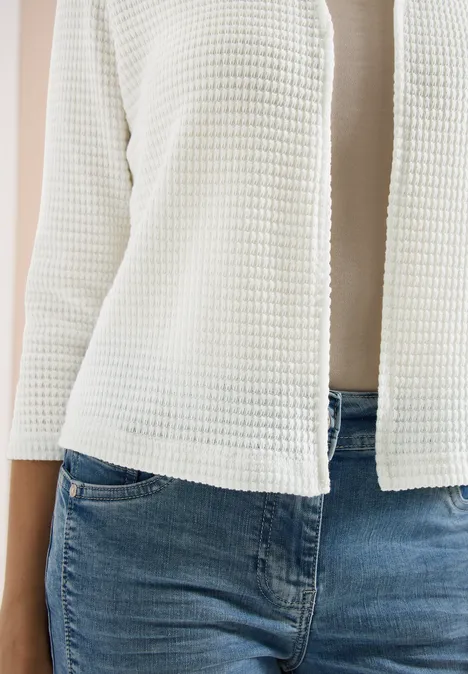 Een vrouw draagt een witte, gestructureerde cardigan over een beige top en een lichtblauwe spijkerbroek.