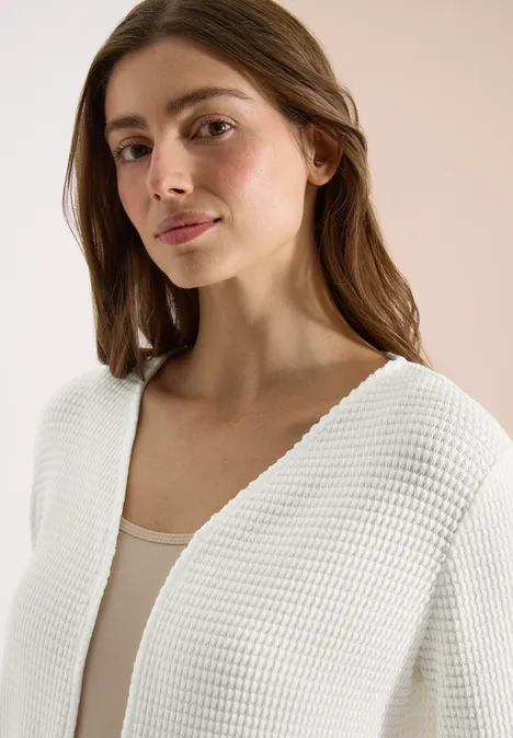 Vrouw draagt een witte grofgebreide vest en een beige hemdje, tegen een ecru achtergrond.