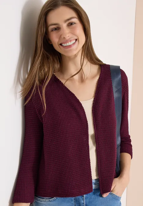 Vrouw draagt bordeaux vest over wit shirt, hand in zak, voor een lichte achtergrond.