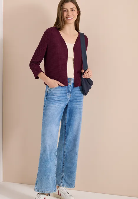 Vrouw in paarse cardigan, beige top en wijde jeans, witte sneakers en donkerblauwe schoudertas voor egale achtergrond.