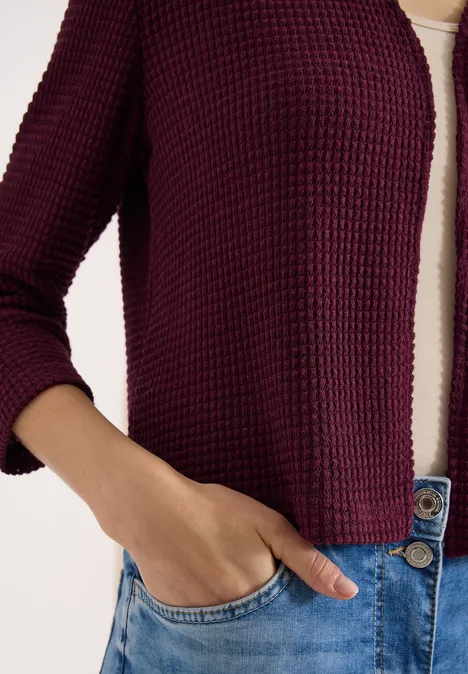 Vrouwelijk bovenlichaam met bordeaux cardigan, wit shirt en blauwe jeans, hand in de zak