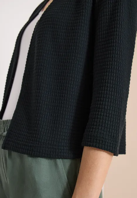 Loszittende, zwarte cardigan met wafelstructuur en driekwartmouwen, gedragen over een wit topje en groene broek.