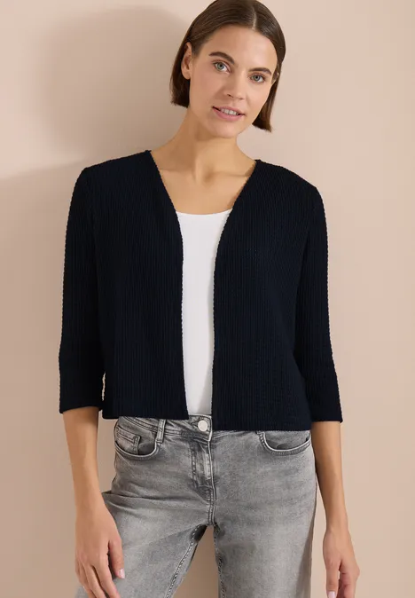 Offene 3/4-Arm Jacke im Ajour-Look universal blue Frau trägt kurze schwarze Strickjacke, weißes Top, graue Jeans vor beigem Hintergrund.