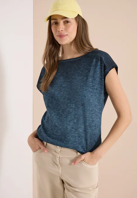 Shirt met boothals in garment-dye look universal blue Vrouw in een blauw T-shirt en beige broek voor een lichte achtergrond, handen in de zakken.