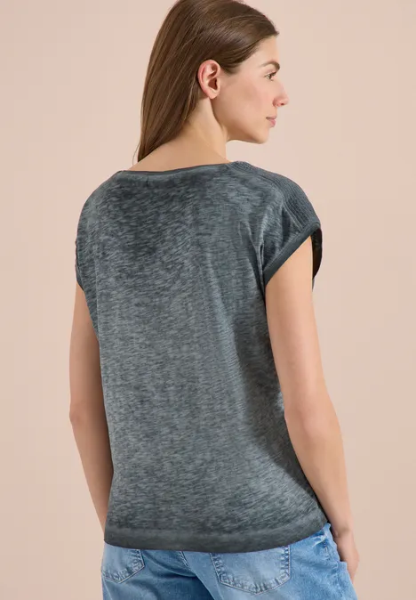 Frau steht von hinten in grauem T-Shirt und Jeans vor hellem, einfarbigem Hintergrund.
