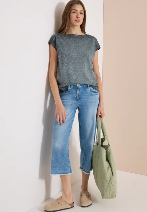 Frau in grauem T-Shirt, hellblauen Cropped-Jeans, beigen Slippern und grünem Steppbeutel vor zweifarbigem Hintergrund.