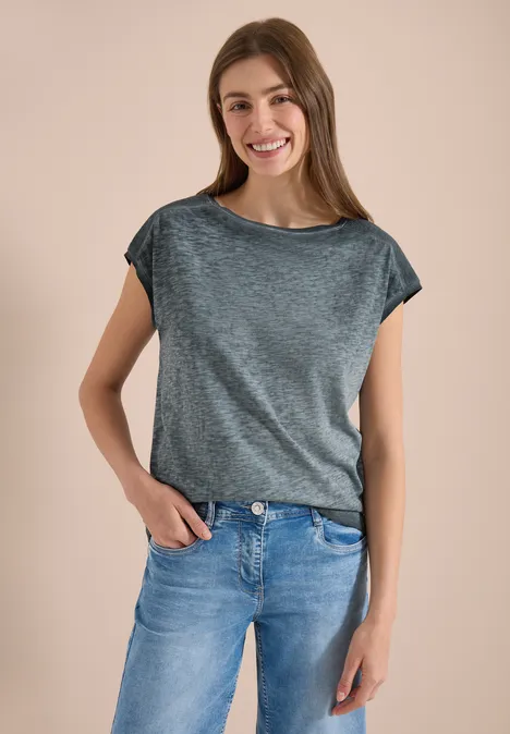 Eine Person trägt ein graues T-Shirt und blaue Jeans, steht vor neutralem Hintergrund.