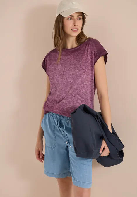 Frau in lila T-Shirt, Jeans-Shorts und weißer Kappe, die eine Jacke in der Hand hält.