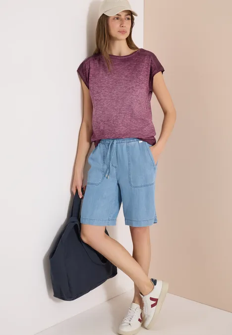 Frau mit lila T-Shirt und hellblauen Shorts, weiße Sneaker, trägt dunklen Rucksack, Gesicht unscharf.