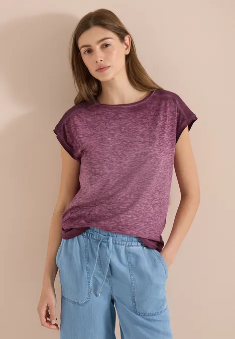 Frau trägt violettes T-Shirt und hellblaue Hose, steht entspannt vor neutralem Hintergrund.