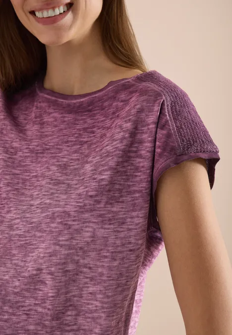 Eine lächelnde Frau in einem violetten kurzärmligen Pullover, nur Oberkörper sichtbar.